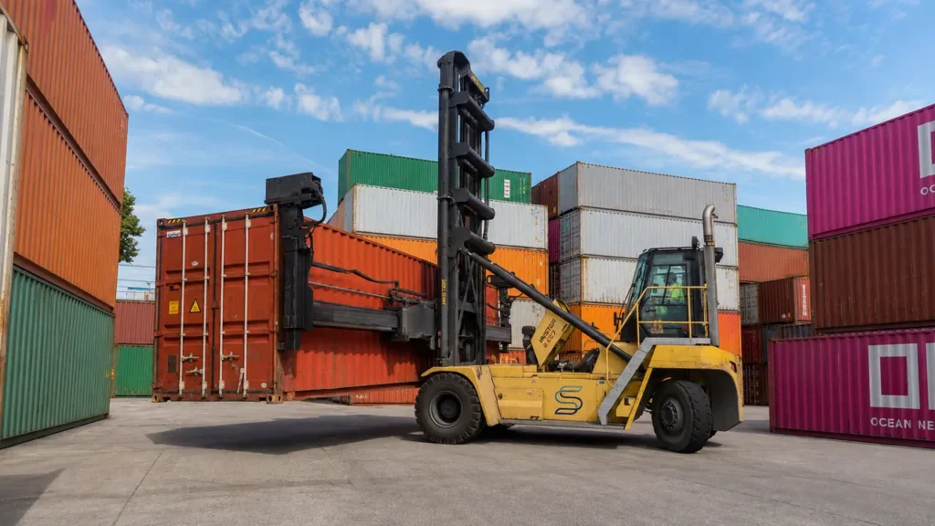 Reach stacker handling a dry shipping container at a depot, illustrating handling and logistics costs for European buyers.