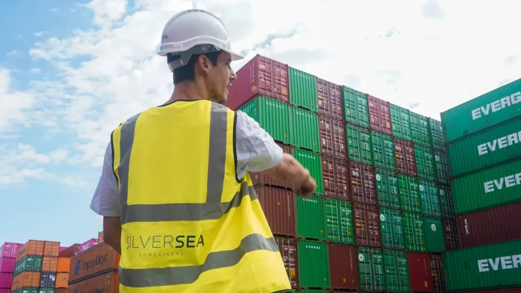 SILVERSEA Containers worker wearing a hard hat and high-visibility vest, pointing towards stacked shipping containers at a depot.