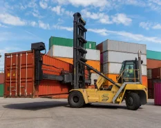 Reach stacker handling a dry shipping container at a depot, illustrating handling and logistics costs for European buyers.